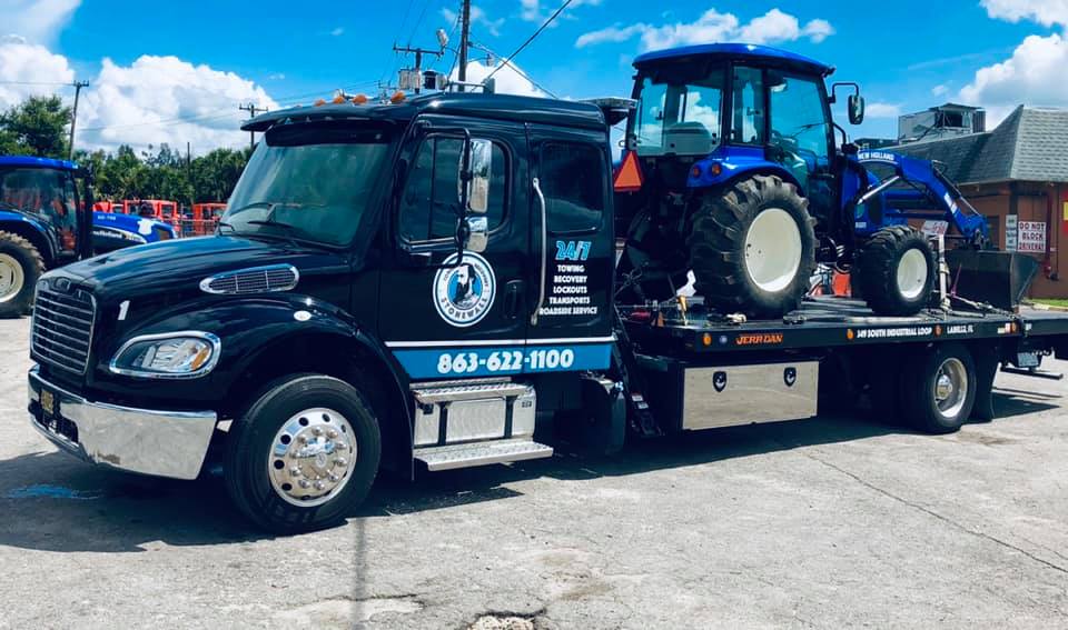 View of one of our flat bed tow trucks with a tractor loaded on it in a parking lot.